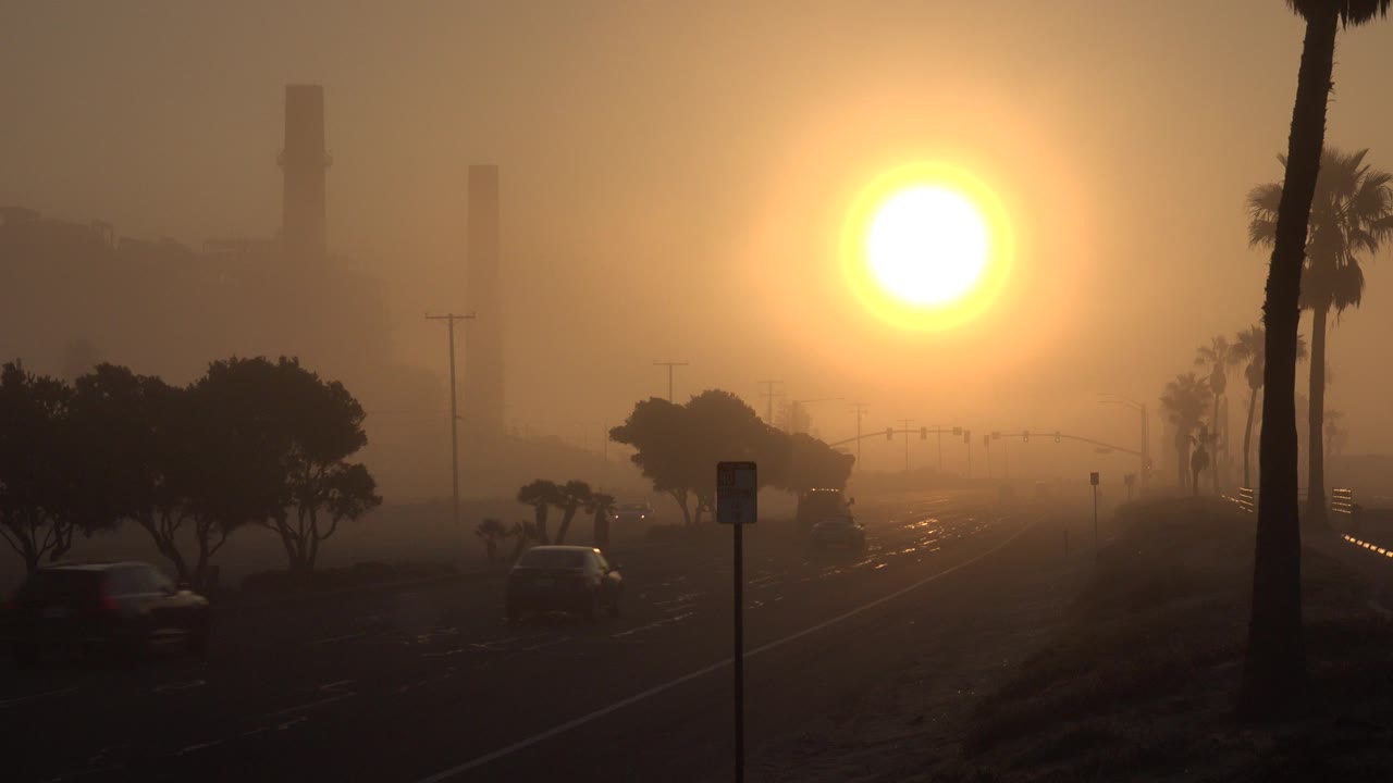 Bright orange sun over a hazy road with palm silhouettes and distant buildings.