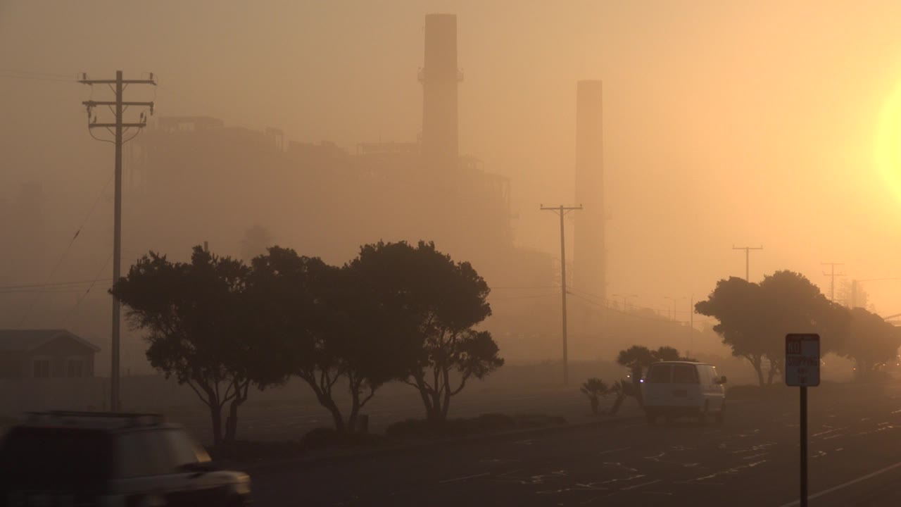 Hazy sunlit road with cars, trees, utility poles, and distant industrial stacks.