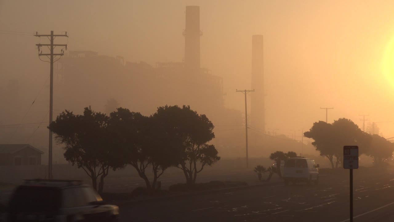 At 0:01 in Huntington Beach, California, a dark car crosses the lower left while distant vehicles continue toward tree silhouettes and softened industrial stacks, with no visible people.