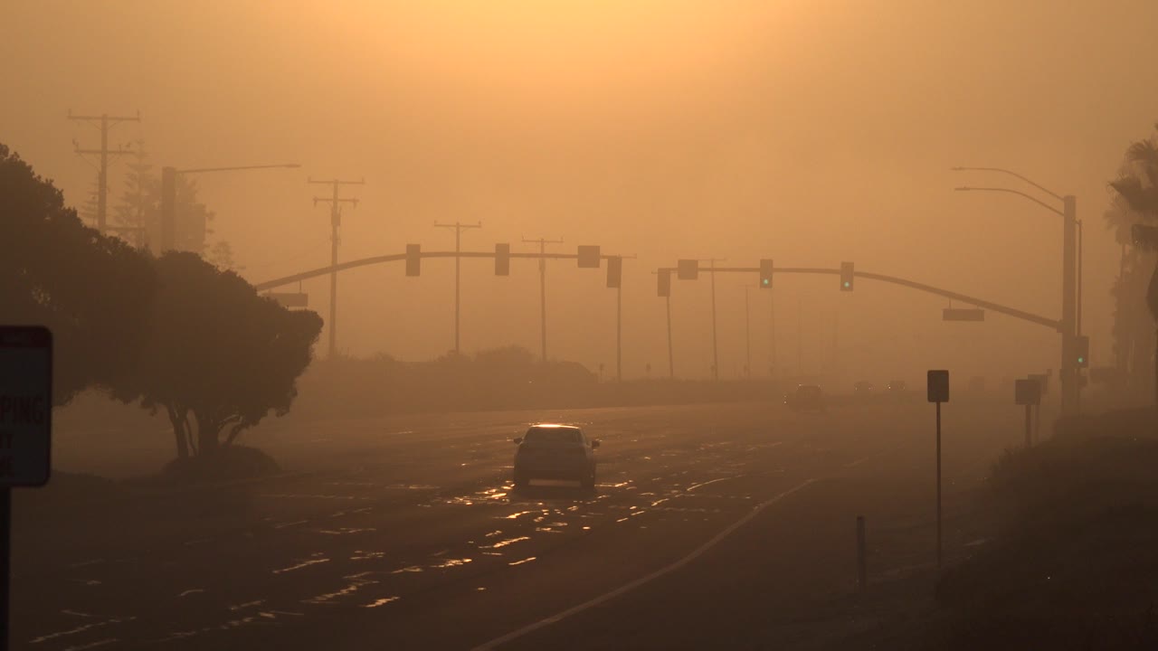 Huntington Beach roadway at orange sunset with overhead signals, sparse traffic, roadside trees, wet pavement reflections, and no visible people.
