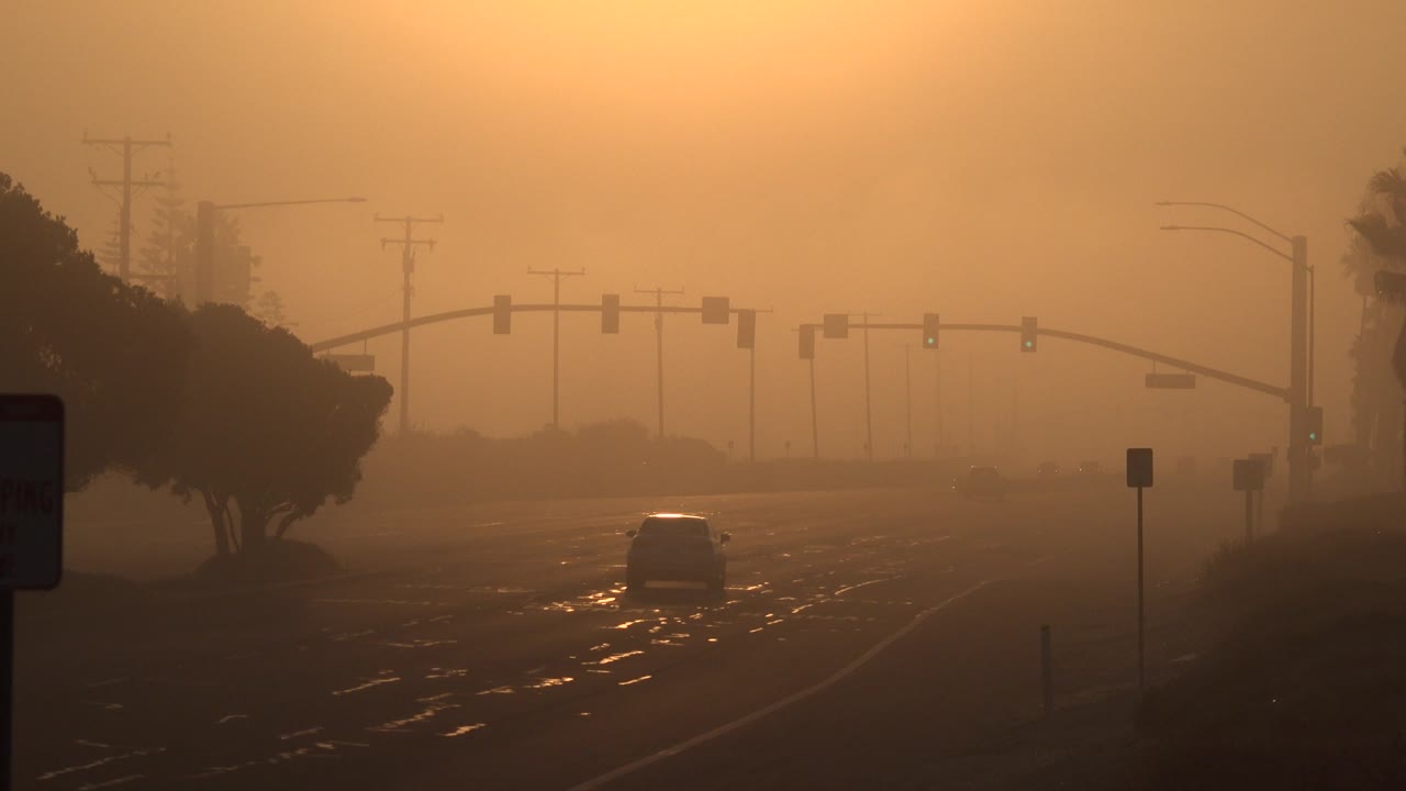 A car moves farther into a hazy Huntington Beach roadway scene, with amber sky, signal arms, utility poles, dark tree silhouettes, and no visible people.