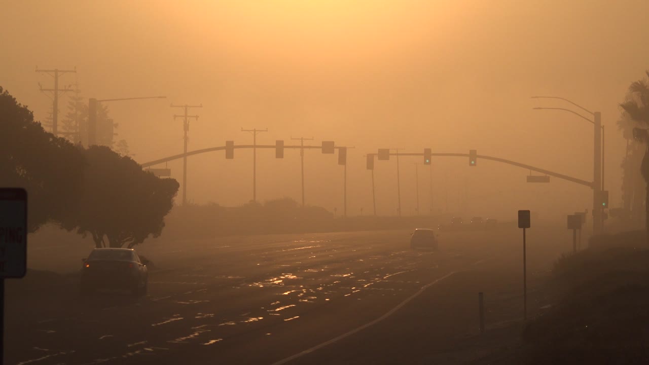 A Huntington Beach road sits under a dense orange sky, with traffic lights ahead, trees at the edges, a car near the left curb, and no visible people.