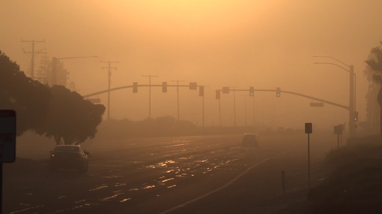 A car moves deeper into a hazy Huntington Beach roadway beneath suspended traffic lights, with dark trees and utility poles silhouetted through the orange light and no visible people.