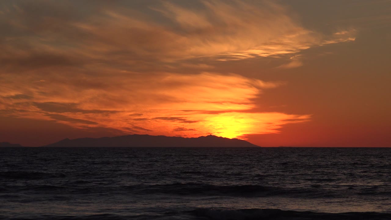 Opening frame of a Huntington Beach sunset with orange light behind distant land, dark choppy ocean water, layered clouds, and no visible people.