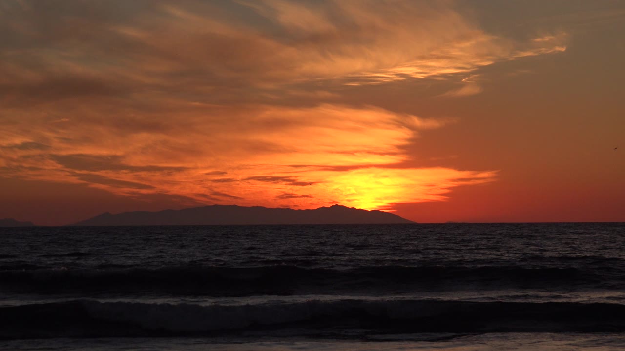 Orange sunset over dark ocean waves and distant landform.