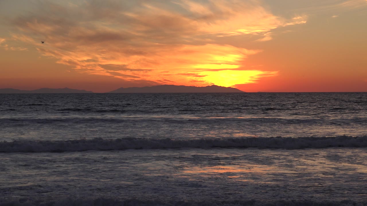Huntington Beach sunset frame with the sun low behind distant land, orange clouds above dark ocean waves, and no visible people.