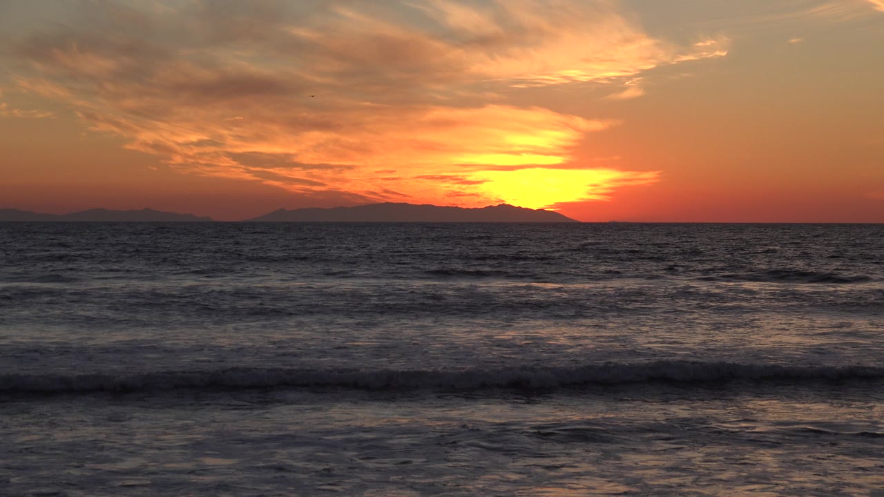Huntington Beach ocean view with the sun glow near the horizon, small waves crossing the frame, distant land in silhouette, and no visible people.