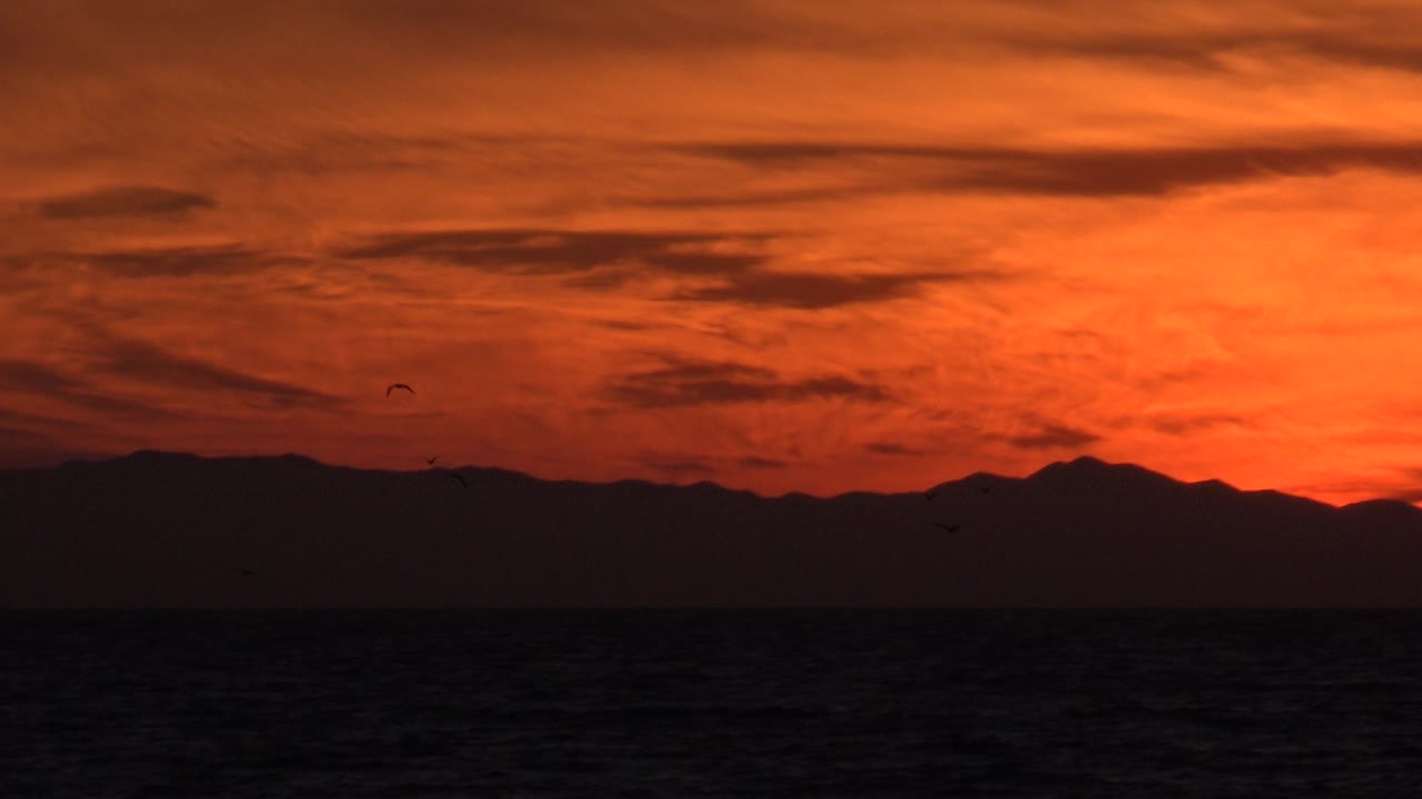 A red-orange Huntington Beach sky sits over a black ocean band and a low mountain ridge, with tiny dark bird silhouettes and no visible people.