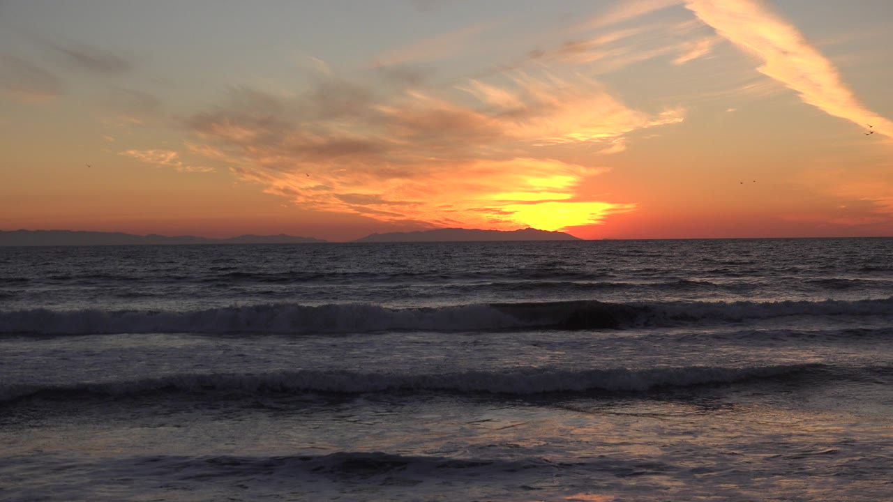 Huntington Beach sunset over dark rolling surf, with a bright yellow sun near the horizon, orange clouds above, distant land silhouettes, and no visible people.