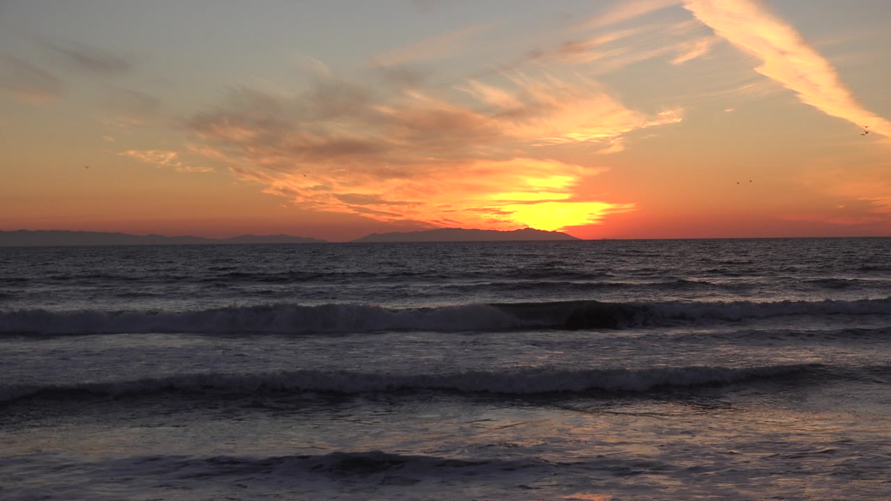 At 0:01, the Huntington Beach horizon remains dark beneath a bright yellow sun, with orange cloud streaks overhead, choppy wave bands below, and no visible people.