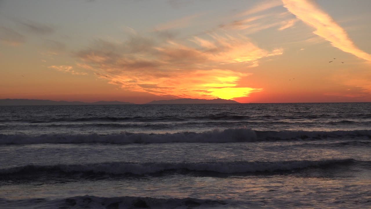 Orange sunset with streaked clouds over breaking waves at the beach.
