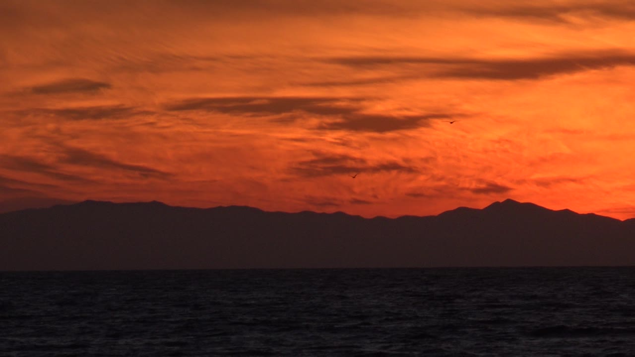 Red sky and silhouetted mountains over the ocean with birds in the distance.