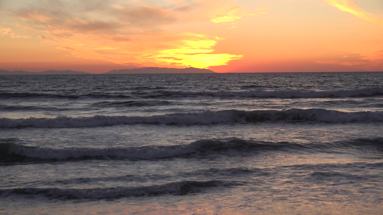 Huntington Beach sunset view with low waves rolling toward shore, distant land on the horizon, warm red-orange sky, and no visible people.