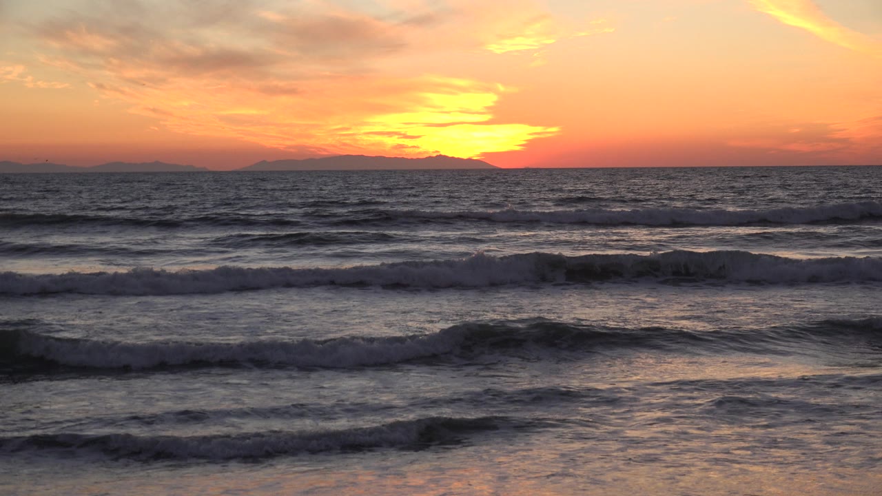 Huntington Beach waves break in layered bands beneath a bright orange sunset glow, with dark distant land along the horizon and no visible people.