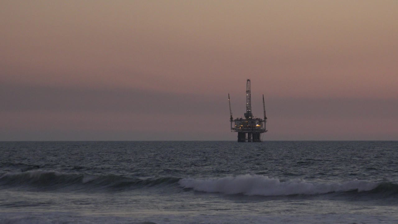 At 0:00 in Huntington Beach, California, dark surf breaks in the foreground while an offshore structure sits near the horizon under a pink-orange sky, with no visible people.