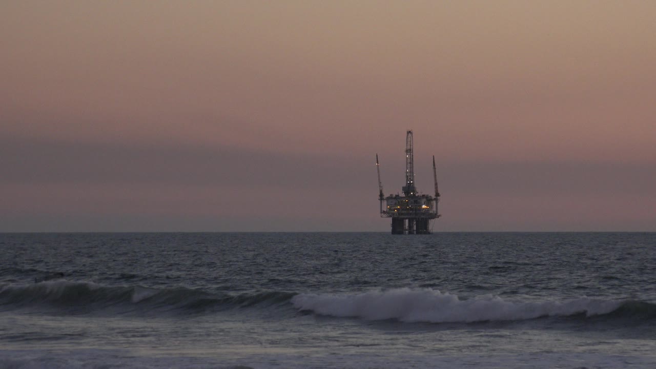 At 0:01 in Huntington Beach, California, a darker wave line crosses the lower frame as the offshore structure remains centered on the horizon beneath muted sunset color, with no visible people.