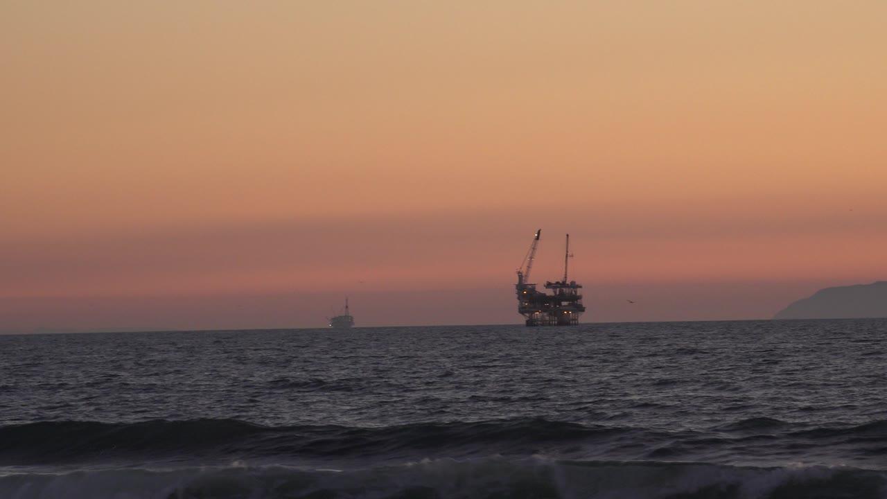 Huntington Beach sunset view with dark waves in the foreground, offshore boat silhouettes near the horizon, a faint landform at right, and no visible people.