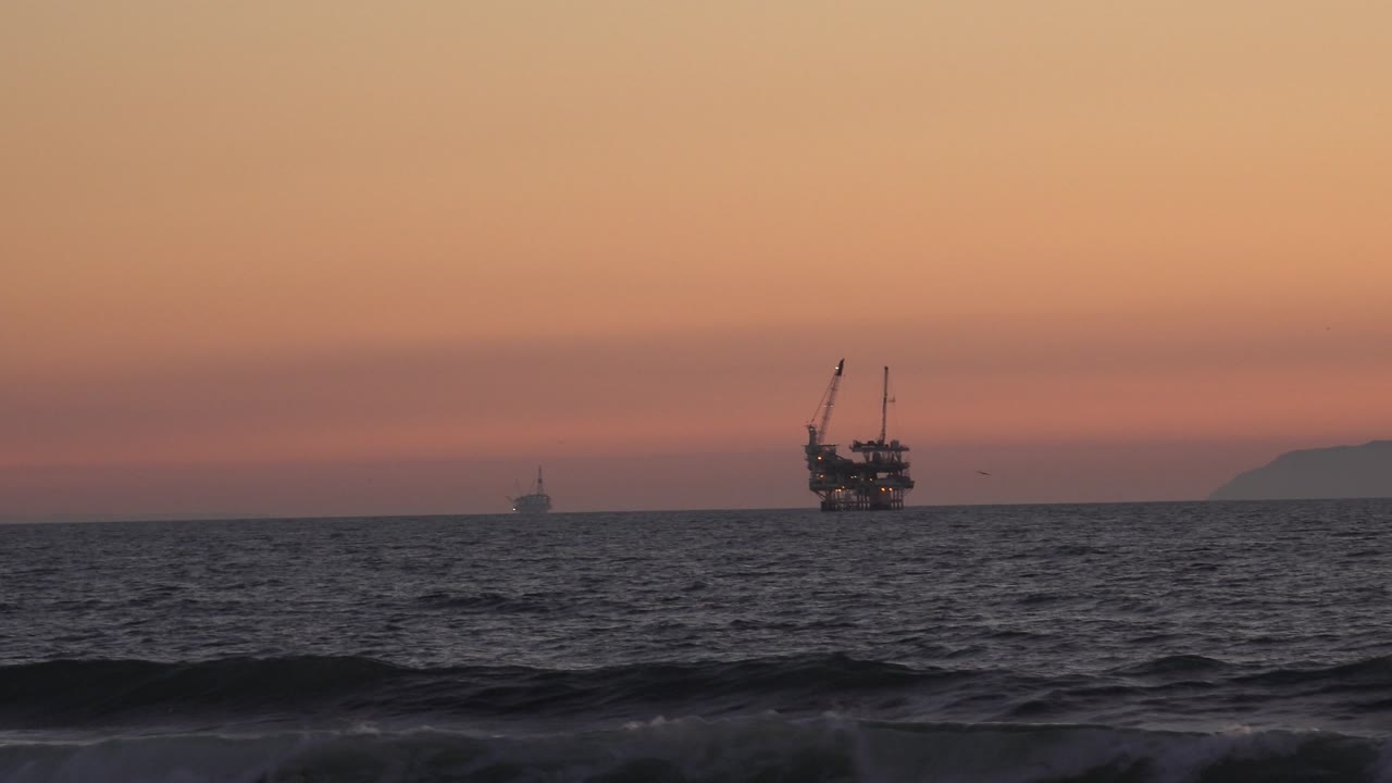 At 0:01, the Huntington Beach horizon holds two dark offshore structures against orange-pink sky while a low wave line crosses the foreground, with no visible people.