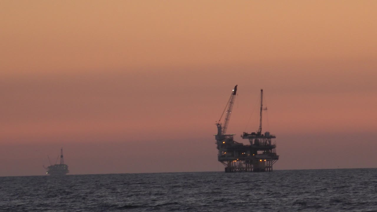Offshore oil platform near Huntington Beach, California, seen across dark ocean water under an orange sky, with a small vessel far left and no visible people.