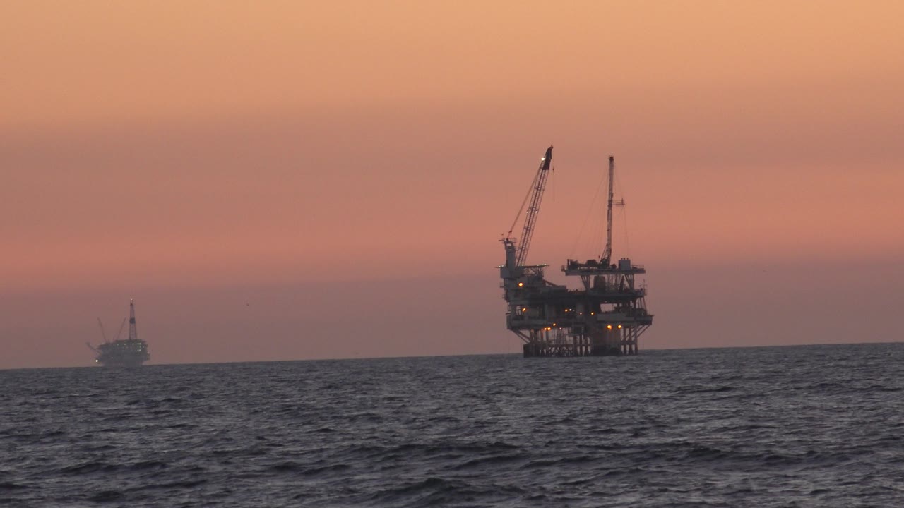 A distant offshore oil platform stands on the right horizon off Huntington Beach, California, with a small vessel far left, dark ocean water in front, peach sunset sky, and no visible people.