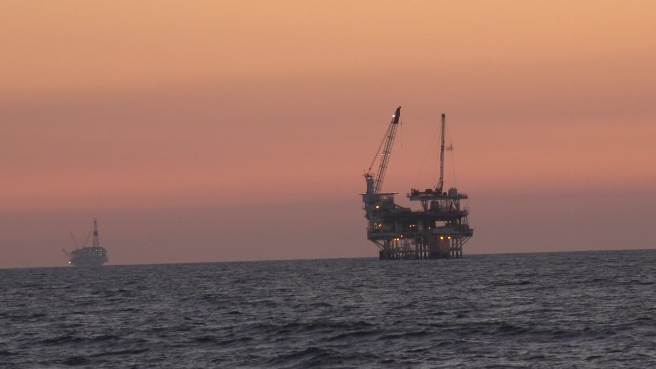The Huntington Beach ocean horizon holds a silhouetted oil platform with two crane arms on the right, a small vessel left of center, rippled water below, and no visible people.