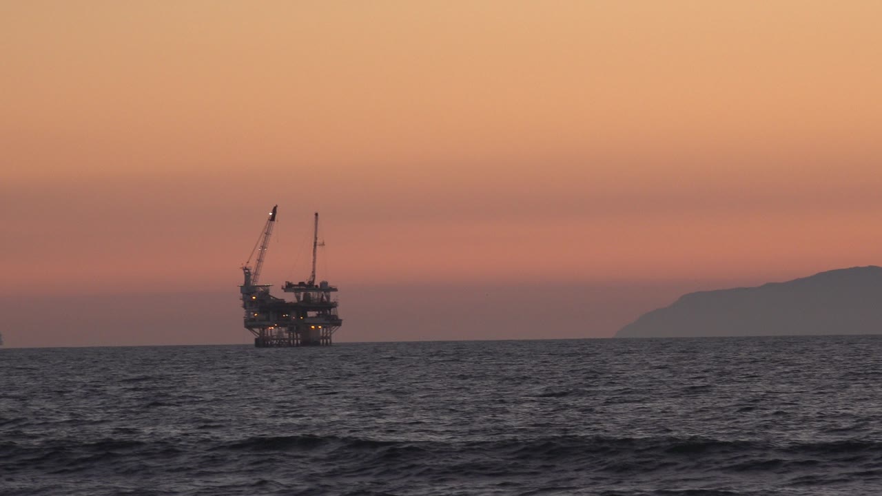 Distant offshore oil platform beyond dark ocean waves at sunset.