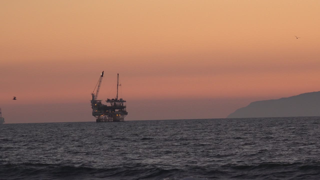 A distant offshore oil platform sits left of center off Huntington Beach, California, with dark waves in the foreground, orange sunset sky above, distant land at right, and no visible people.