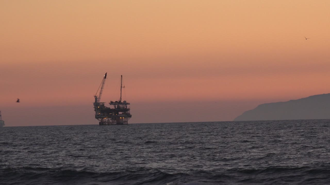 The oil platform remains near the left side of the Huntington Beach horizon, with rippled dark water below, a peach-orange sky above, a landform at right, and no visible people.