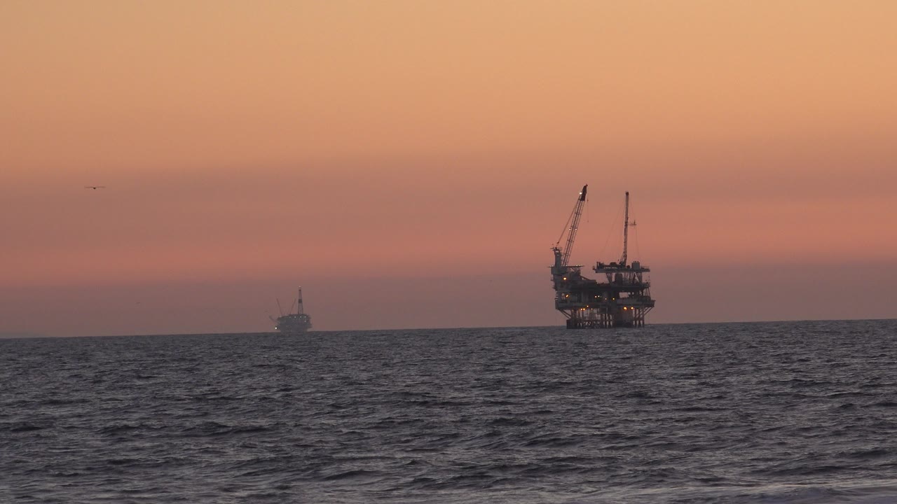 Huntington Beach sunset view across dark ocean waves toward a large offshore silhouette with cranes and a smaller distant shape, with no visible people.