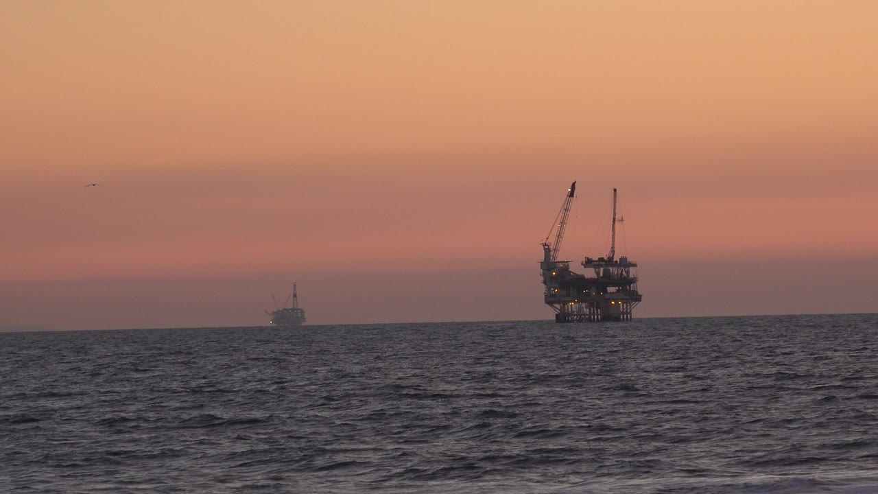 Huntington Beach ocean horizon at sunset, showing choppy foreground water, a large crane-topped offshore structure, a smaller distant vessel or structure, and no visible people.