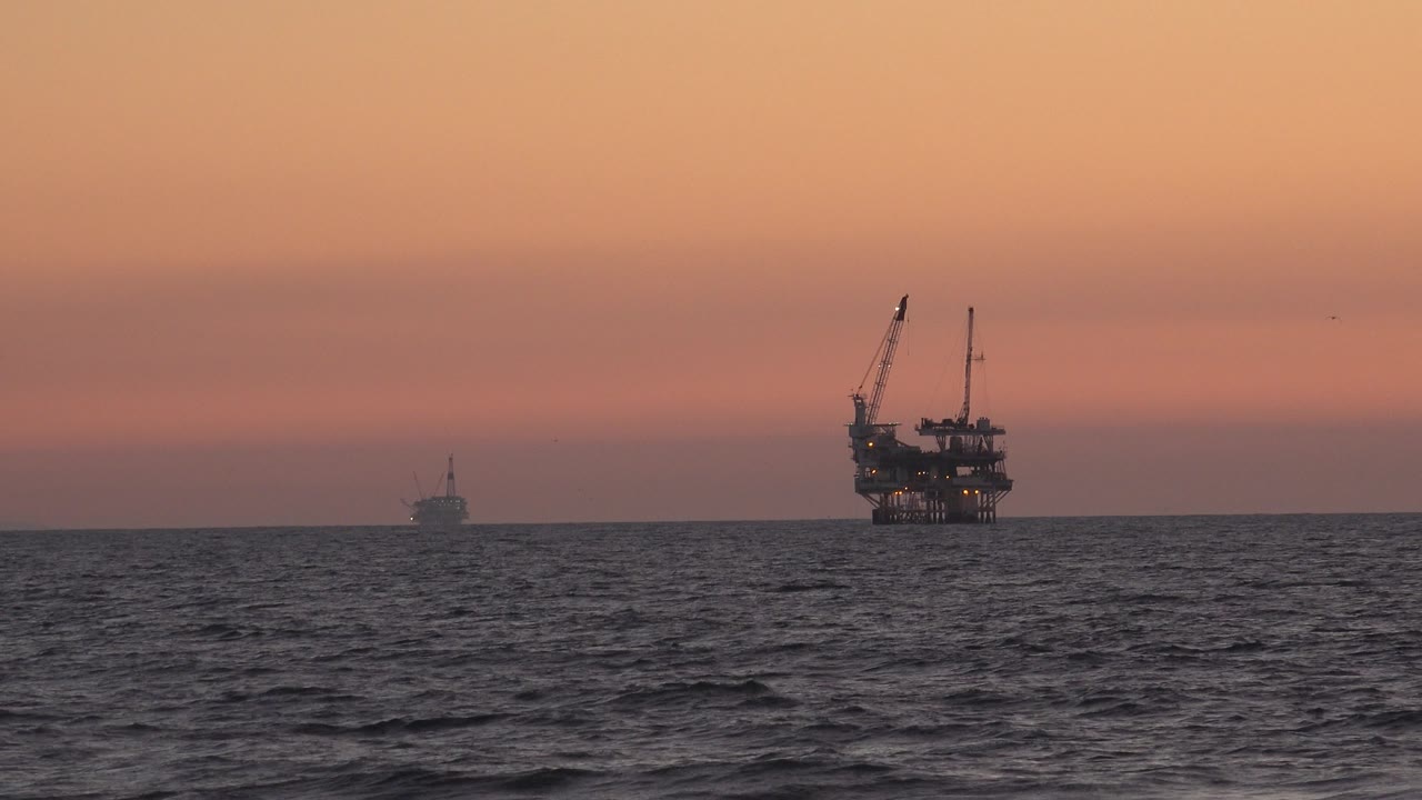 At Huntington Beach, California, dark ocean water sits below a red-pink sunset sky with a small distant silhouette on the left and a larger offshore structure to the right, with no visible people.