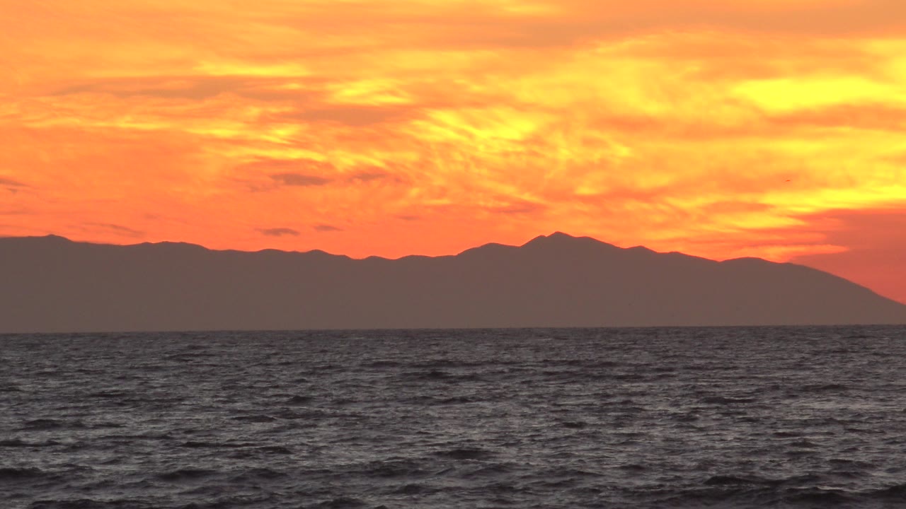 At 0:00 in Huntington Beach, California, dark ocean water sits below an orange sunset sky and a distant mountain silhouette, with no visible people.
