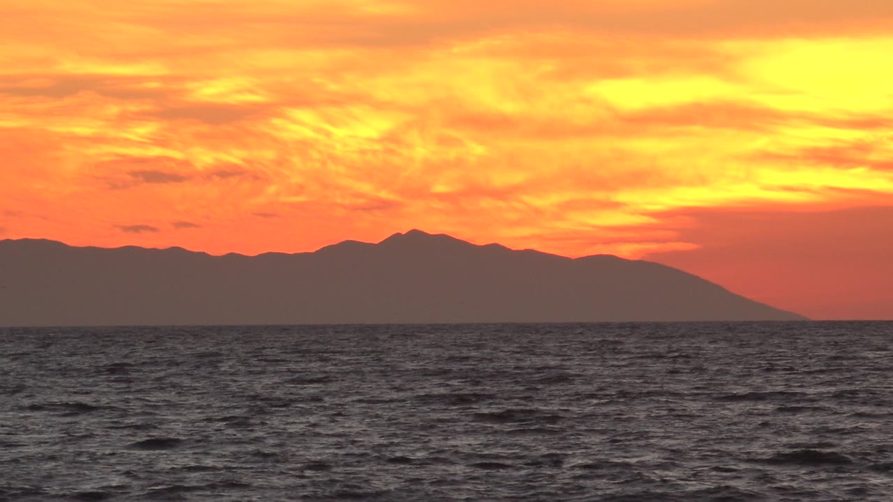 Opening frame of a Huntington Beach sunset with rippled ocean in front, a dark land mass across the horizon, orange clouds above, and no visible people.