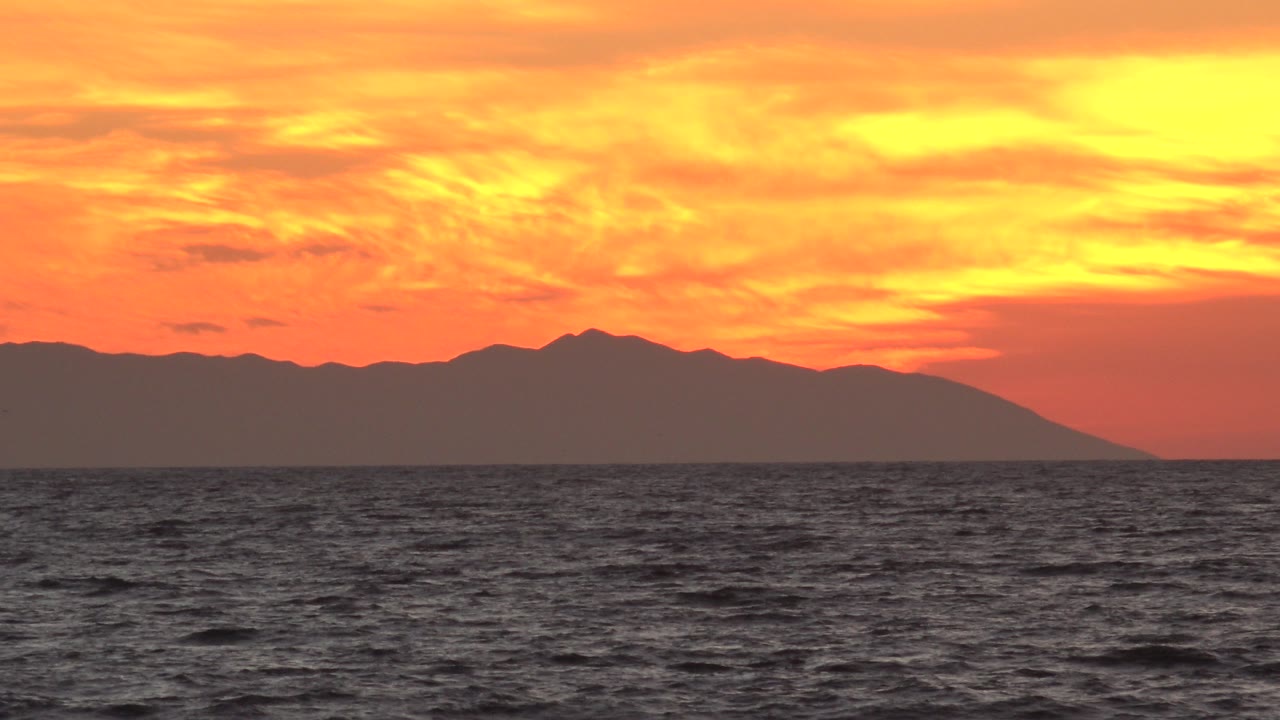 Second frame from Huntington Beach showing a wide band of dark water, a silhouetted land mass centered on the horizon, glowing orange cloud cover, and no visible people.