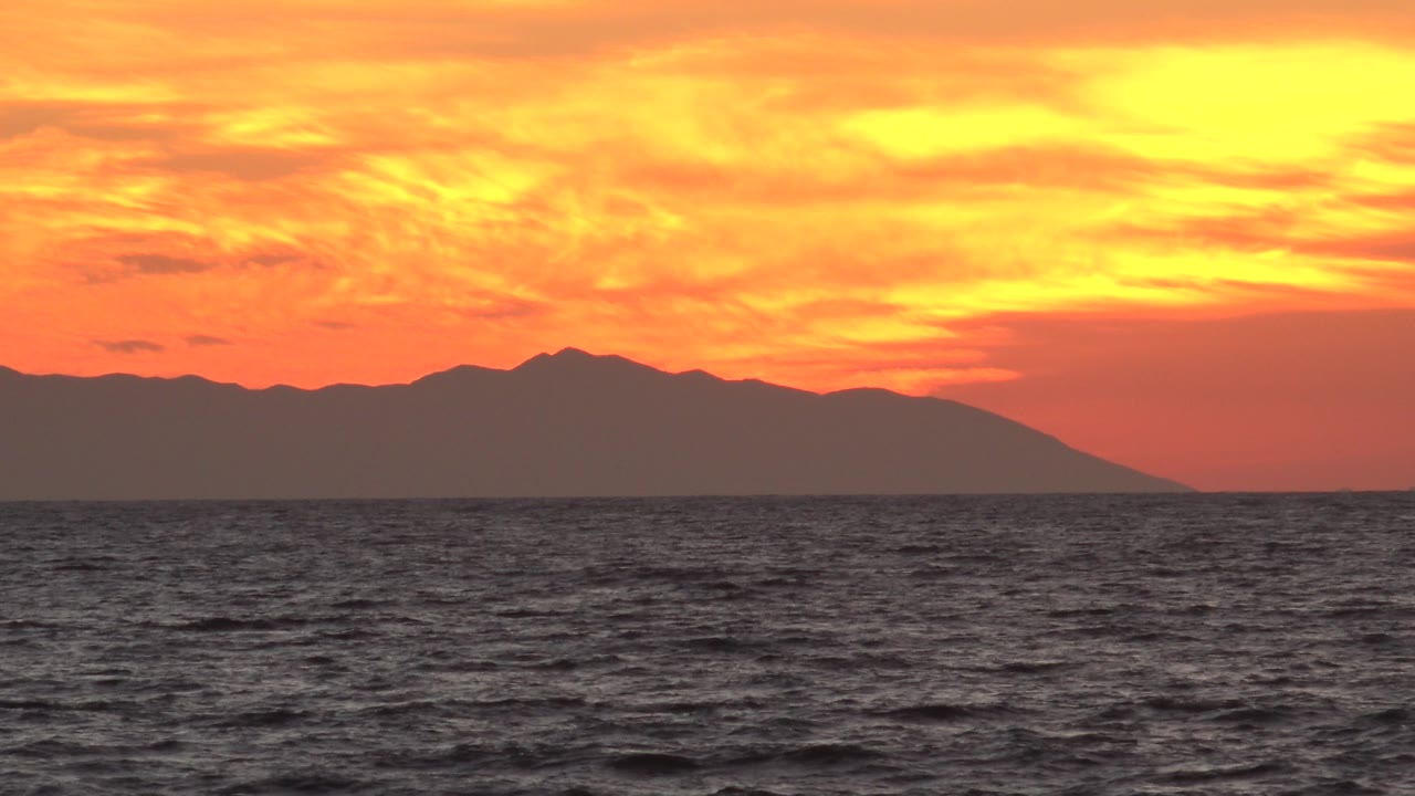 Sunset clouds over ocean with a dark mountain-like land mass.