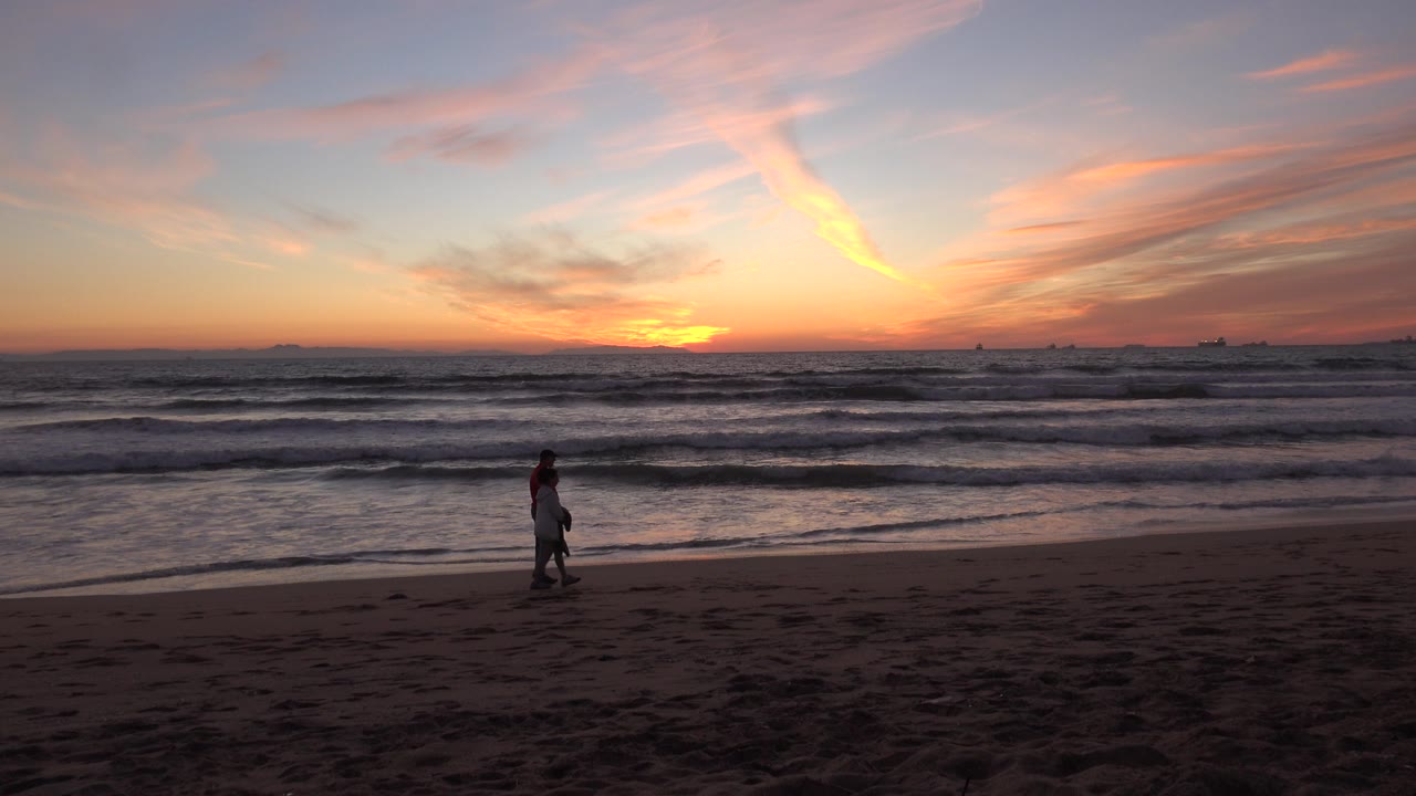 Couple walking on a beach at sunset with waves behind them.