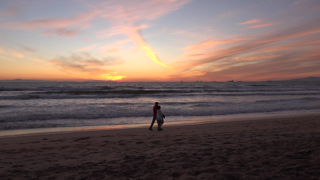 At Huntington Beach, California, a couple walks near the shoreline under an orange sunset, with dark sand in front and rolling waves behind them.