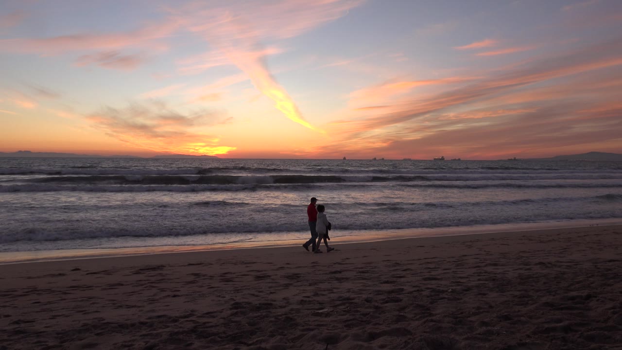 Couple on the sand with waves and pink sunset clouds behind them.