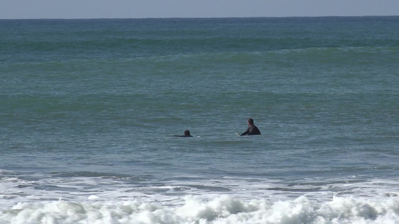 Wide Huntington Beach frame with two distant surfers in dark wetsuits beyond foamy shore break under sunny light.