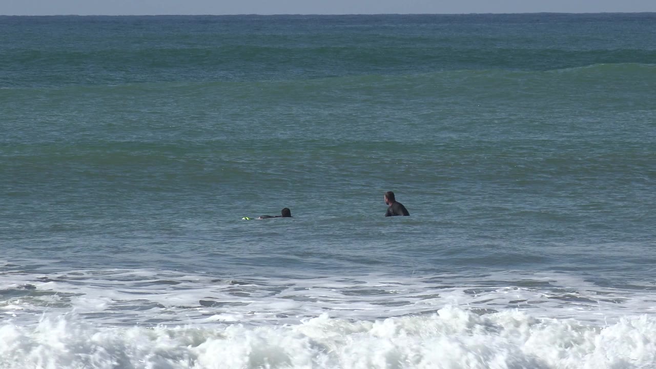 Huntington Beach surf scene with a paddling surfer and a seated surfer separated in the midground beyond the foamy break.