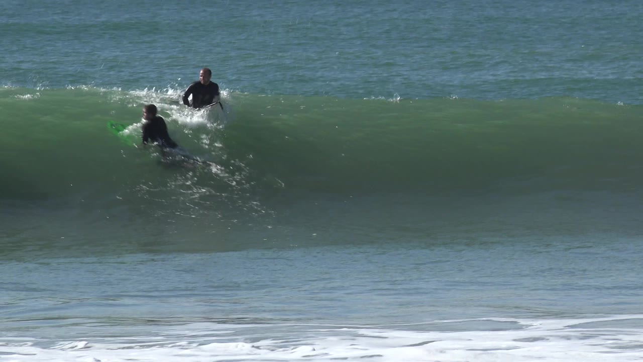 At Huntington Beach, California, two surfers remain separated across a dark green wave face as the ocean horizon stays level behind them.