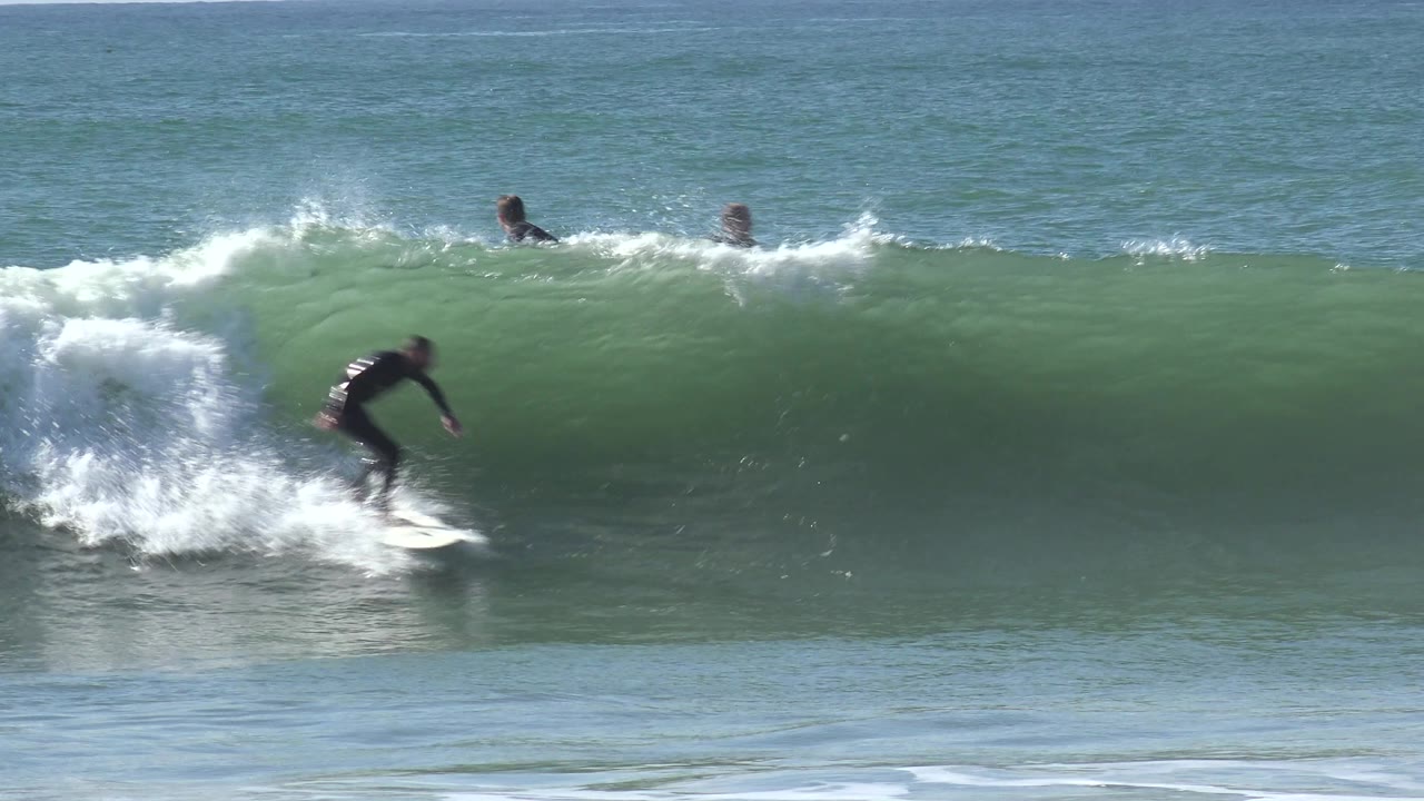 At Huntington Beach, California, a surfer in a black wetsuit rides down the face of a green wave while whitewater breaks behind and other surfers are small in the background.