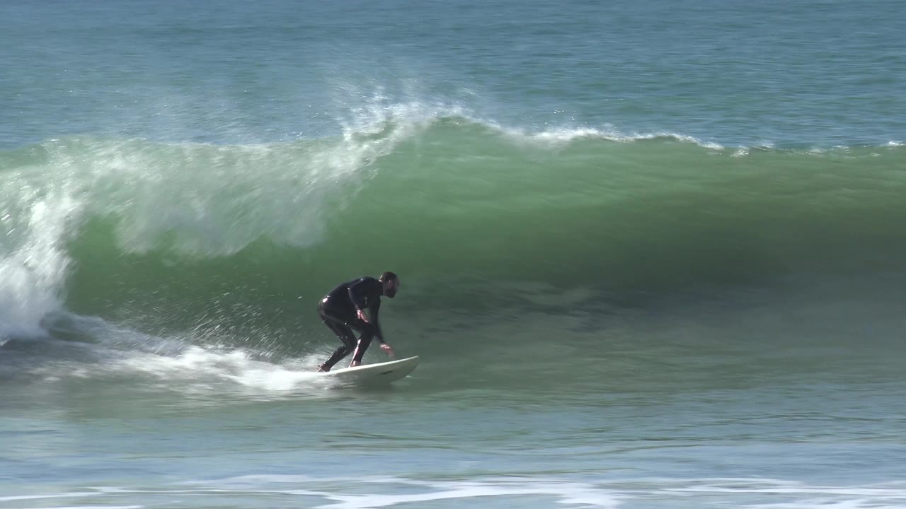 At Huntington Beach, California, the surfer rides low on a white board along a smooth green wave face, with breaking foam trailing to the left and open water above.