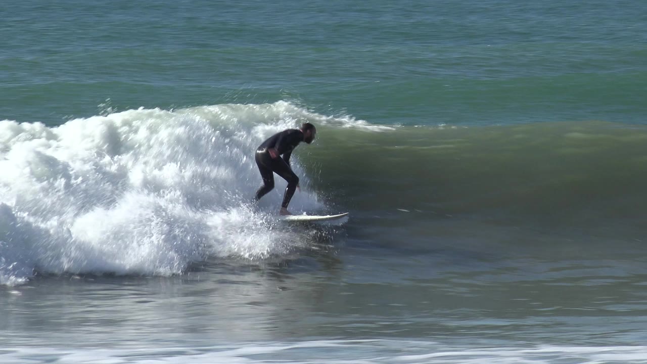 A surfer in a dark wetsuit rides a white surfboard across breaking whitewater at Huntington Beach, California, with green-blue ocean behind.