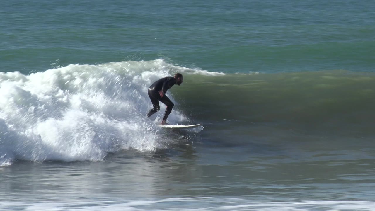 The surfer crouches low on a white surfboard at Huntington Beach, California, with a curling wave and bright spray rising beside the board.