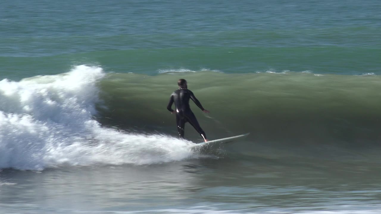 A surfer on a white board rides near the right edge of the frame at Huntington Beach, California, with a smooth green wave face behind and white spray trailing left.