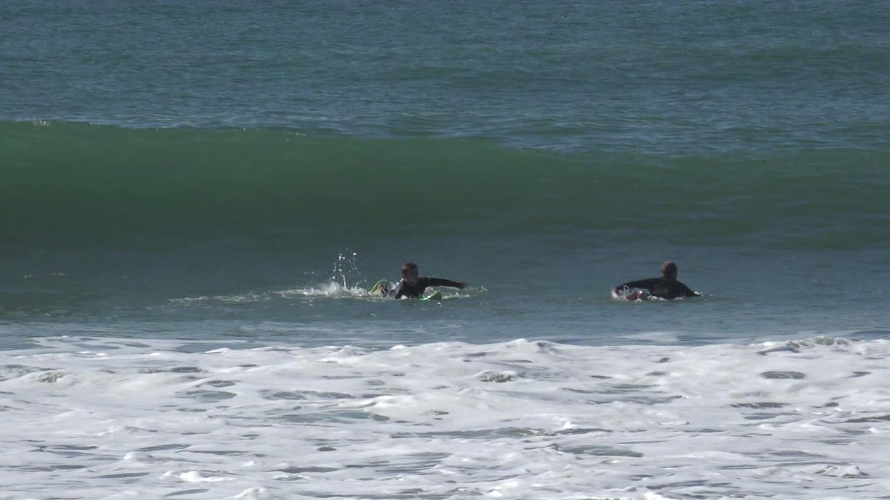 Two surfers in dark wetsuits paddle in the ocean at Huntington Beach as a green wave rises behind them and white foam washes across the foreground.