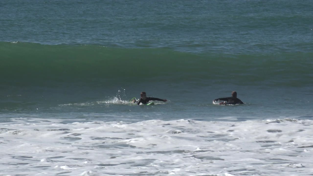The surfers remain low on their boards at Huntington Beach while the wave face steepens behind them and foam rolls through the lower part of the frame.