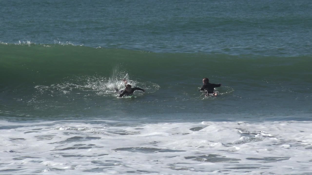 A surfer at Huntington Beach kicks up spray while starting onto the wave, with a second surfer nearby and a clean green wall of water behind them.