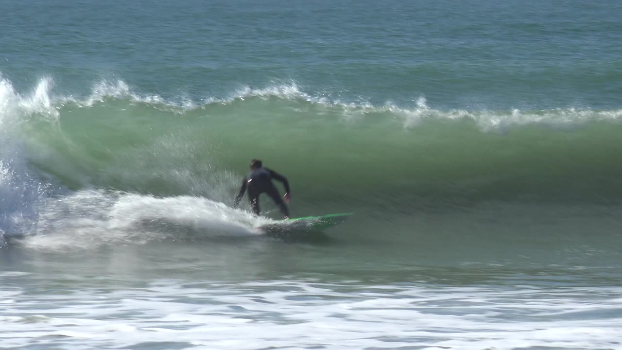 A surfer in a black wetsuit rides a green board along a green-blue breaking wave at Huntington Beach, California, with white foam spreading in front.